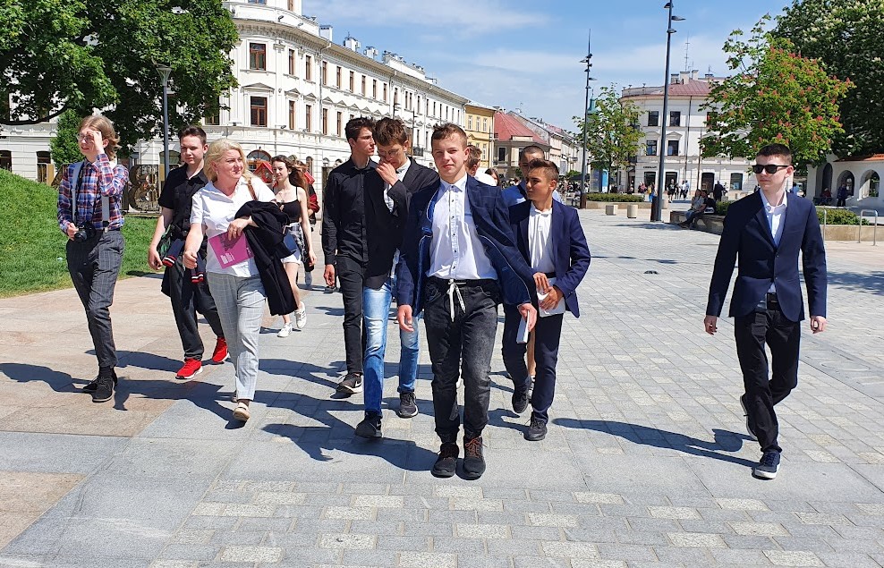 Na zdjęciu grupa młodych ludzi wraz nauczycielką idących chodnikiem przez miasto.